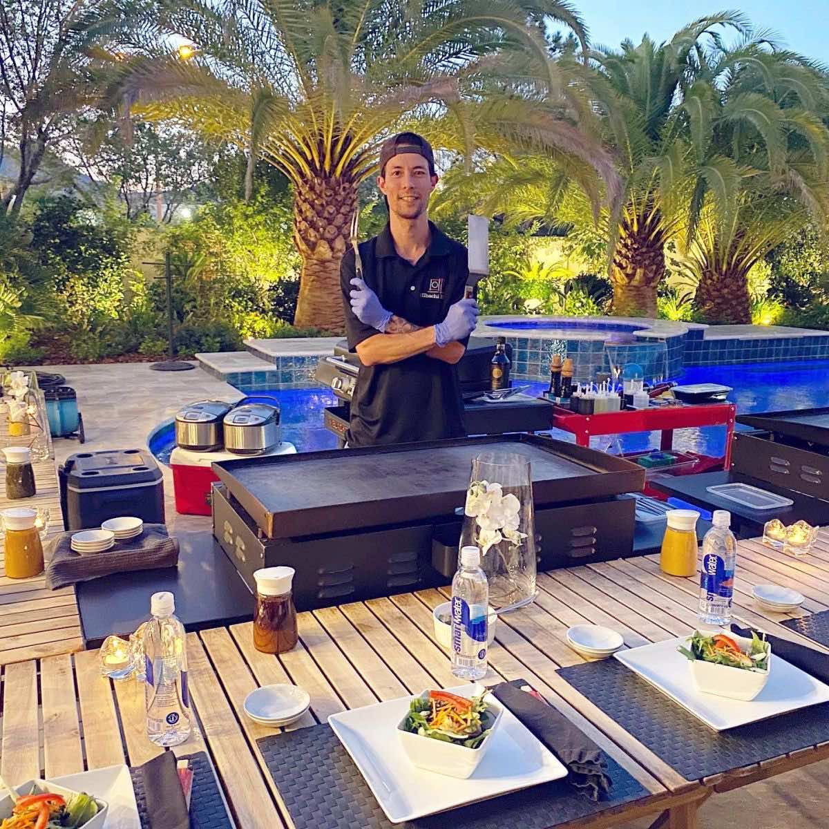 Chef Koma, arms crossed with spatula and knife, behind his griddle before a poolside dinner party